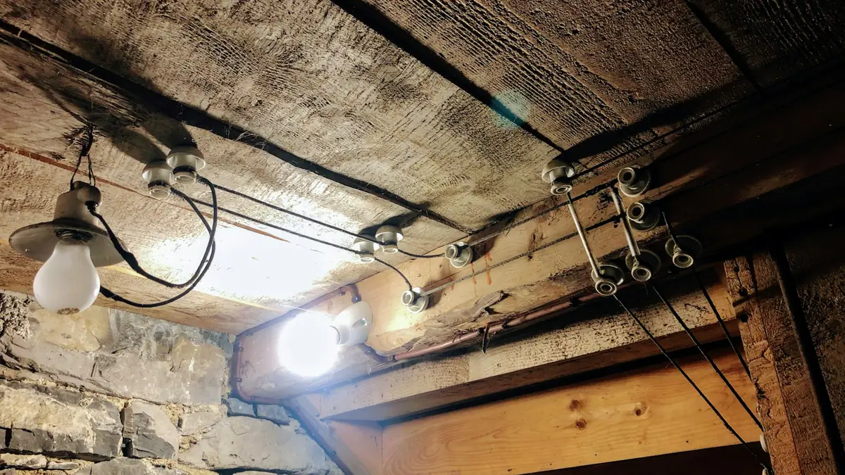 An old basement ceiling with exposed joists, wiring and a bare light bulb - Leak Detection in Beachmont